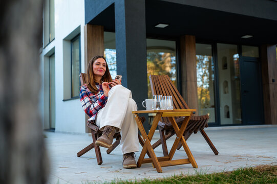 Young woman sits on a wooden patio chair at a modern house, smiling while scrolling her smartphone, relaxing outdoors and enjoying peaceful, casual leisure time and digital connection