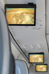 Interior second-class aircraft cabin with seated unrecognizable passengers and shared overhead screens displaying flight information during the journey and the destination "Ahmed Ben Bella Airport"