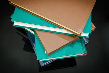 Stos książek na błyszczącej powierzchni. Widok z góry na stos kilku książek z twardymi okładkami, w kolorach brązowym, turkusowym i granatowym, ułożonych na lśniącej, ciemnej  powierzchni  © PIOTR D