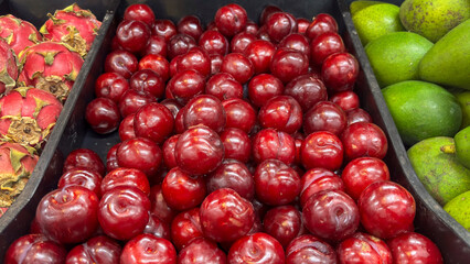 Close-up of bulk red plums, arranged in a black crate for sale at a market or fair. The fruit has a smooth peel and intense color. Surrounded by dragon fruit and avocados.