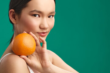 Asian woman holds an orange near her shoulder, smiling gently against a vivid green backdrop,...