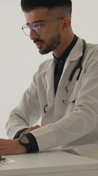 Vertical video. Young male healthcare worker in a white coat and stethoscope is focused on typing on a laptop at a desk in a contemporary hospital environment, fully engaged in his work