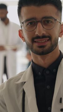 Vertical video. Handsome male doctor in a white coat and glasses smiling confidently at camera while diverse clinic team works behind him