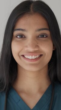 Vertical video. Young indian female doctor in scrubs with stethoscope, smiling confidently in a clean white background headshot