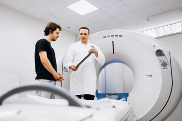 Medical professional in white coat discussing CT scan results with male patient in black shirt, showcasing advanced imaging technology in a clinical environment