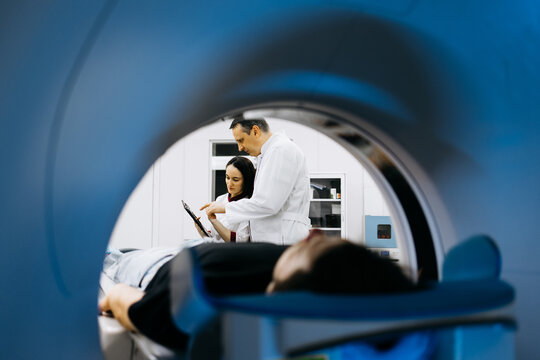 Medical professionals, a male doctor and female technician, are reviewing patient data near a CT scanner, emphasizing advanced imaging technology and patient care in a clinical environment