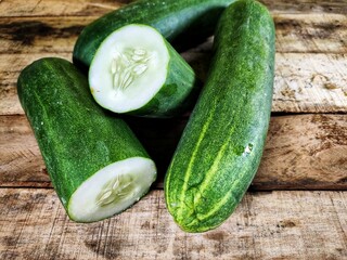 A fresh green cucumber is cut in half on a small, rustic wooden cutting board, revealing the seeds and juicy interior, with other whole cucumbers in the background.