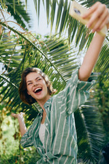 Bright, joyful portrait of a woman outdoors taking a selfie among lush palm leaves, wearing a striped shirt and radiating summer energy.