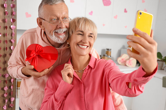 Happy mature couple with gift box and mobile taking selfie in kitchen at home. Valentine's Day celebration