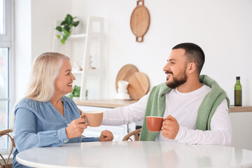 Young man and his mother with cups of tea hugging at table in kitchen