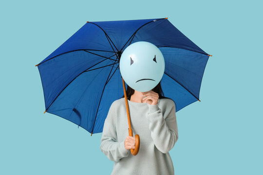 Young woman holding umbrella and balloon with sad face on blue background. Blue Monday concept - Powered by Adobe
