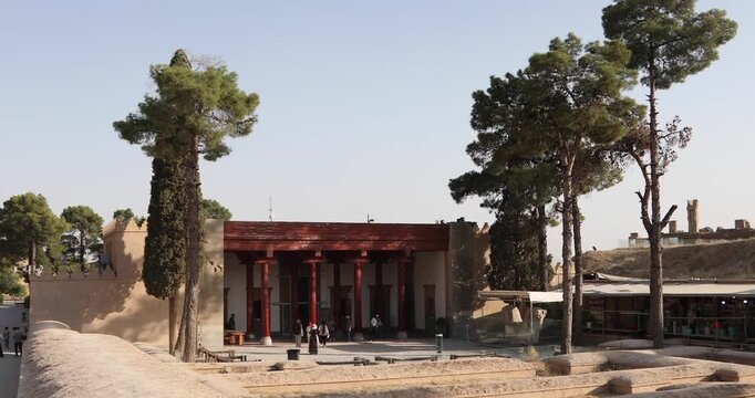 Entrance pavilion at Persepolis, Iran, with restored red columns, tall pine trees, and a few visitors near site museum and bazaar stalls, all in warm afternoon light at the UNESCO World Heritage ruins