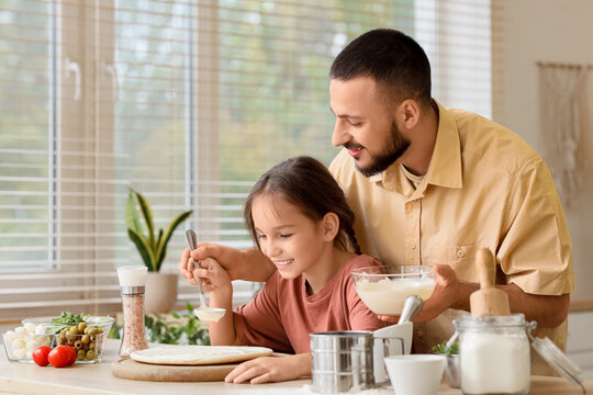 Father with his daughter spreading sauce on pizza dough at table in kitchen