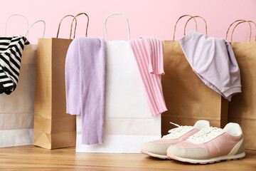 Paper shopping bags with stylish sweaters, t-shirt and sneakers on wooden table against pink wall