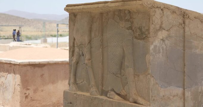 Close-up of a weathered Achaemenid bas-relief at Pasargadae, its carved legs and robes cracked by time. Two visitors stand soft-focused near a fence in the arid landscape.