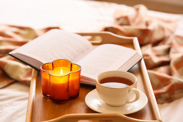 Tray with burning candle, book and cup of tea on bed in bedroom, closeup