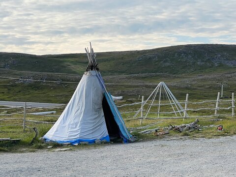Sami raindeer herder tent called lavvu on a side of a road with tundra landscape in backround. Uncovered carcass of another lavvu next to it.