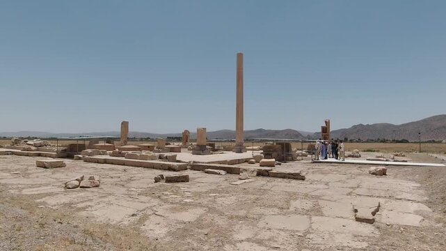 Wide tracking view of Pasargadae palace remains in midday sun: a solitary Achaemenid column rising among stone foundations as a small tour group explores via walkway.