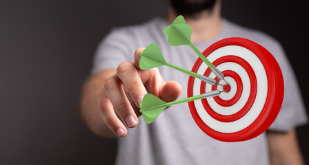 Close-up of a hand pointing at a dartboard with three darts hitting the center. This image...