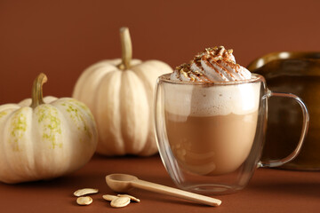 Glass cup of hot pumpkin latte with whipped cream and seeds on brown background