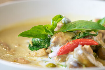 Close up to Green Curry with Chicken garnished on top with pepper and basil was served with white bowl put on wicker cream color placemat.