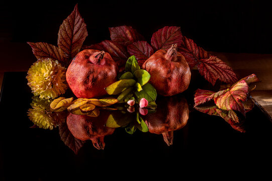 Two pomegranates with autumn leaves, flower, and small buds arranged creatively on shiny black surface. Harvest season concept. Rosh Hashanah. In low key. - Powered by Adobe
