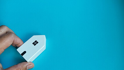 Minimalist concept image showing a hand gently holding a small wooden house on a blue background, symbolizing home ownership, safety, real estate, family care, and housing security.