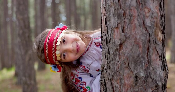 Beautiful Smiling Girl Peeking Behind Tree in Forest Wearing Traditional Ethnic Embroidered Bulgarian Folk Costume symbol of Bulgaria and Independence