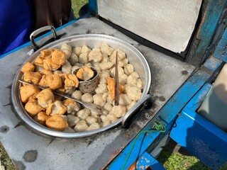 Traditional Indonesian street food cart serving hot fried meatballs and tofu, close-up detail under sunny outdoor light.