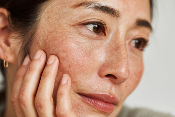Asian woman with freckles touching her face while looking thoughtfully