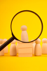 Magnifying glass highlighting a standout candidate among wooden figures, representing recruitment, talent selection and the search for the best fit.
