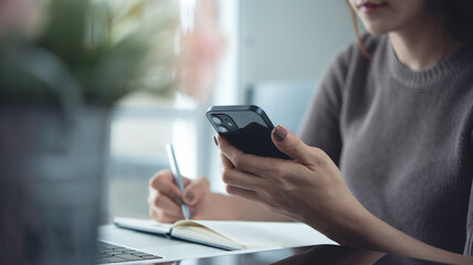 Business woman using smartphone and taking notes on notebook while working on laptop computer, online business, work from home office. Student studying online class, e-learning
