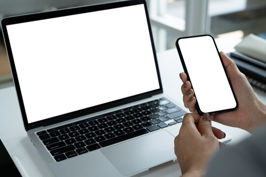 Cell phone and laptop white screen mockup, Woman hand holding mobile phone with blank screen while working on laptop at office. Business woman using smartphone, mockup for social media marketing