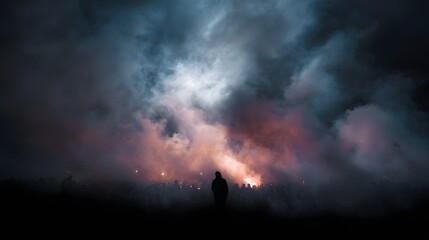 A solitary person stands before a dense crowd at dusk enveloped in dramatic smoke illuminated by orange and white lights