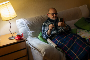 Elderly Man Relaxing on Sofa At Home Using Smartphone With Blanket and Warm Lamp Light