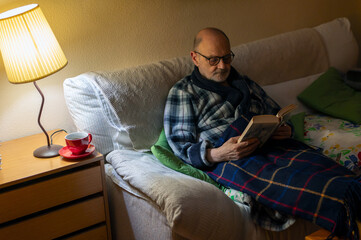 Older Man Reading Book on Sofa With Blanket, Lamp, and Cozy Home Vibe