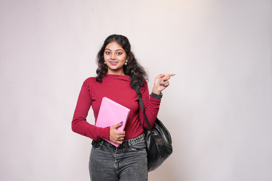 Happy indian female college student with backpack and books pointing at blank space for advertisement isolated on white background. Young asian gen z adult girl looking at camera. Admission open