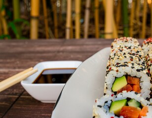Delicious sushi rolls with sesame seeds and fresh vegetables, served with soy sauce and chopsticks on a wooden table with bamboo background