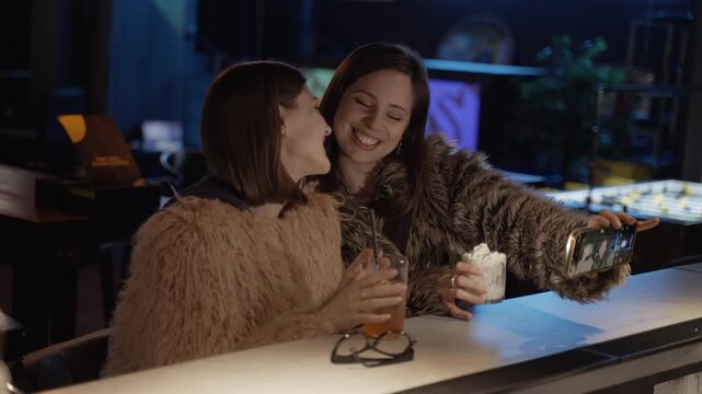Two young women enjoying cocktails and taking selfies with a smartphone flash at a modern bar in the evening