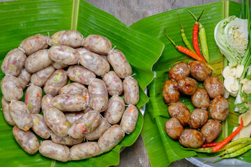 Raw and grilled Thai Isaan sausages side by side on banana leaf with chili, garlic and cabbage, showing local Thai food culture.