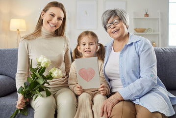 Three happy women generation posing together on sofa, looking gladfully, have happy festive mood during 8 March, hold bouquet of flowers and greeting card. Woman Day celebration concept