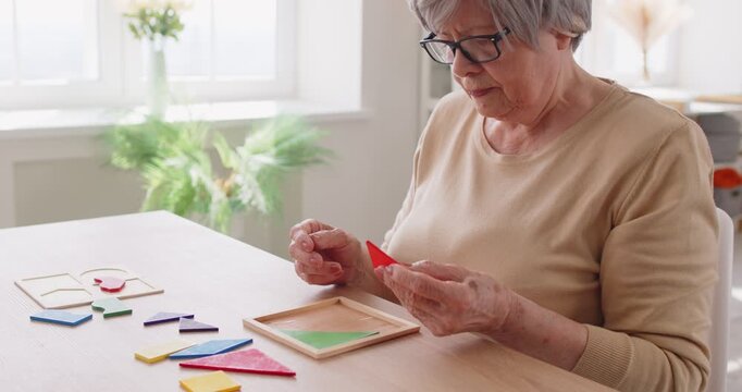 Focused senior woman with gray hair and glasses sitting at wooden table and solving colorful puzzle game. Serious elderly lady assembling pieces, engaging in cognitive activity for memory stimulation.
