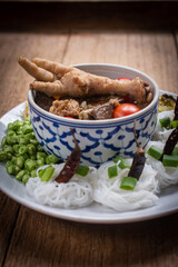 Khanom Jeen Nam Ngiao, a traditional northern Thai noodle soup served with chicken feet, tomatoes and fresh vegetables on a wooden table. Ingredients such as cabbage, long beans and dried chilies.