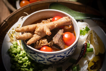Khanom Jeen Nam Ngiao, a traditional northern Thai noodle soup served with chicken feet, tomatoes and fresh vegetables on a wooden table. Ingredients such as cabbage, long beans and dried chilies.