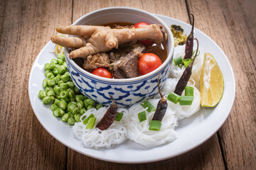 Khanom Jeen Nam Ngiao, a traditional northern Thai noodle soup served with chicken feet, tomatoes and fresh vegetables on a wooden table. Ingredients such as cabbage, long beans and dried chilies.