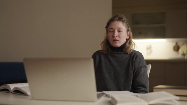 Tired young woman with cerebral palsy finding it hard to stay awake while studying late at night, eventually dozing off in front of laptop and textbook in softly lit home setting, slow motion.