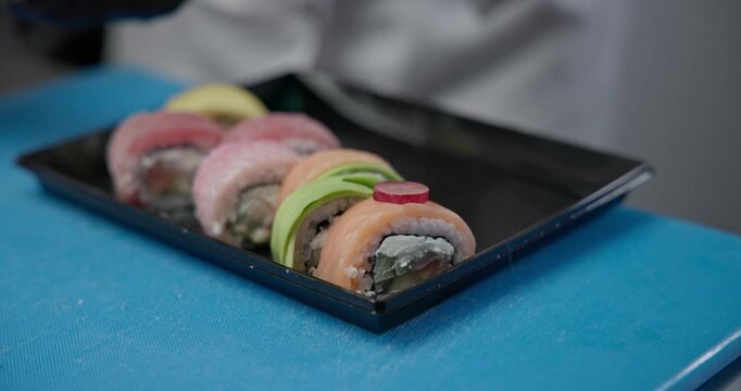 A chef in black gloves decorates sushi rolls with tuna, salmon, and mango with pieces of grapes on a plastic delivery tray.