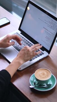 Hands of a woman programmer typing code on a laptop keyboard while sitting at a wooden table in a coffee shop with a cappuccino and a smartphone, working remotely on a new project