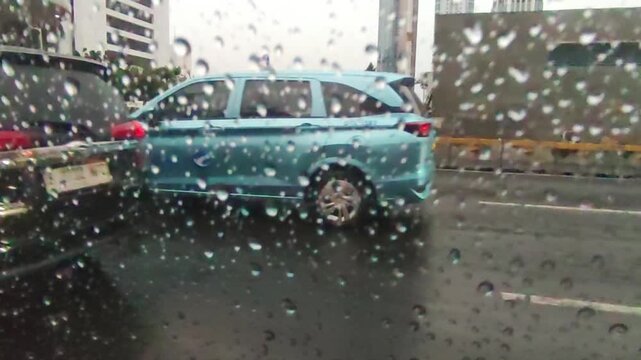 Teal minivan driving on wet city street during heavy rain, creating a blurred and atmospheric scene with reflections and movement, evoking a sense of urban life in inclement weather.