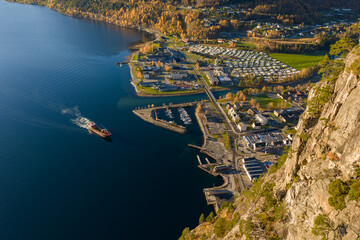 A breathtaking aerial view of a coastal town beside a calm blue fjord, showing a red ship sailing away, marina docks, houses, autumn trees, and steep rocky cliffs bathed in golden sunlight.